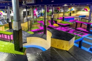 Large indoor trampoline park with colorful trampolines, angled ramps, and soft padded obstacles for jumping and freestyle activities