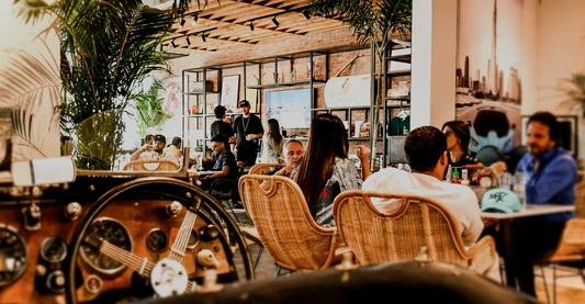 Indoor view of The CARS Café at Dubai Autodrome with people dining, cozy wicker seating, racing-themed wall decor, and part of a vintage car dashboard in the foreground.