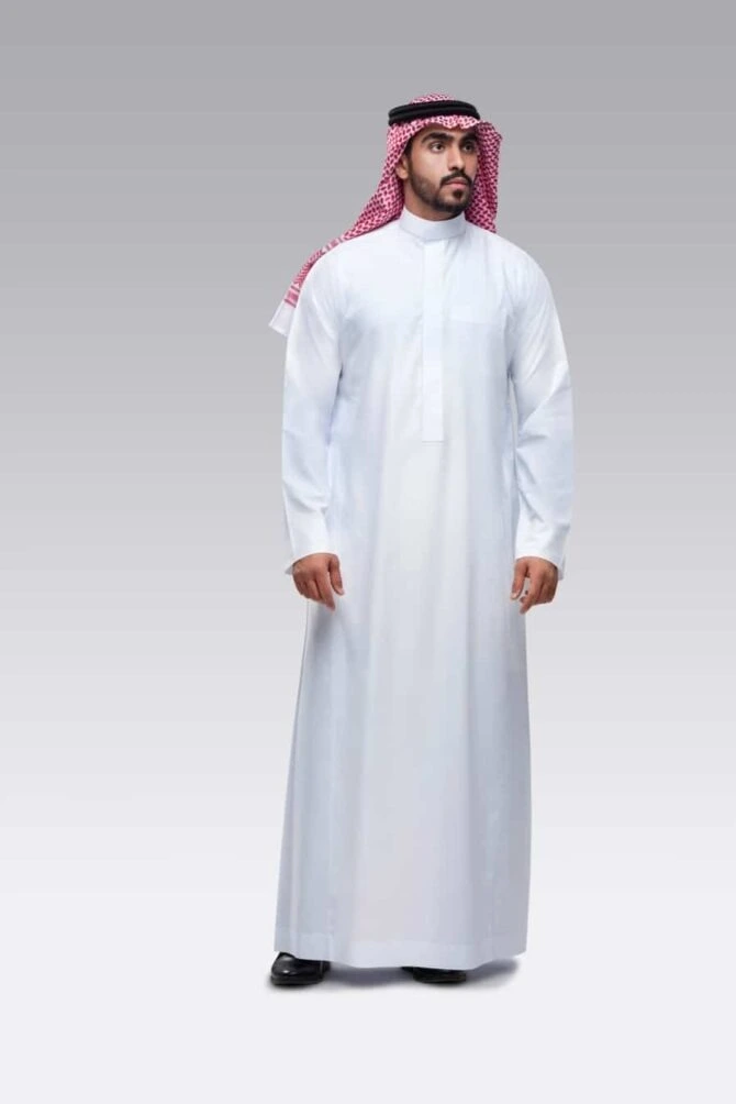 A man models a classic white kandura with a red-and-white checked ghutra and a black agal, standing against a gray slanted background.