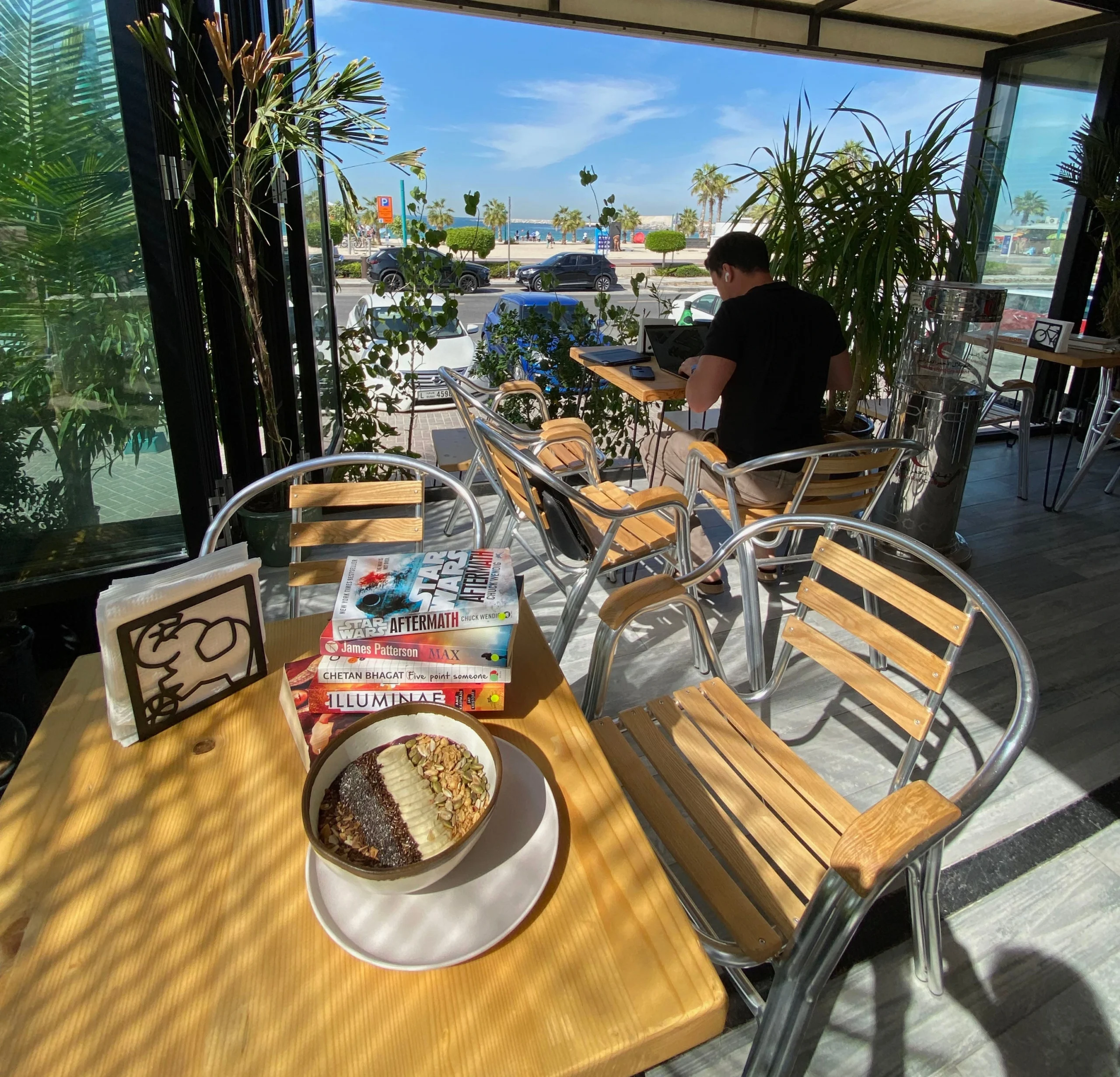 A stack of books sits next to a smoothie bowl on a wooden table with a calm view of the beach in the background.