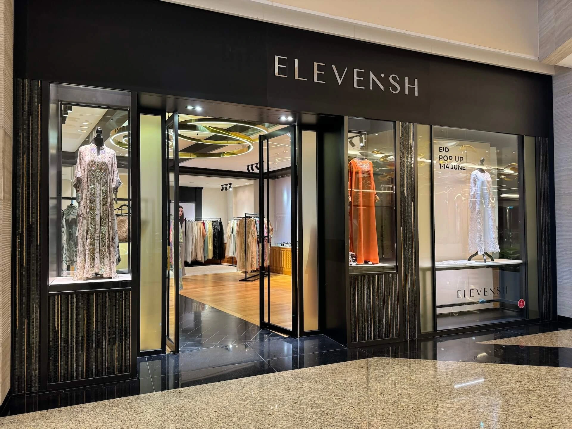 The front of Elevenish Boutique in a shopping mall features elegant dresses displayed on mannequins.