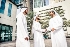 Three Emirati men dressed in white kanduras and black agals are smiling and greeting each other outside in a modern area of Dubai.