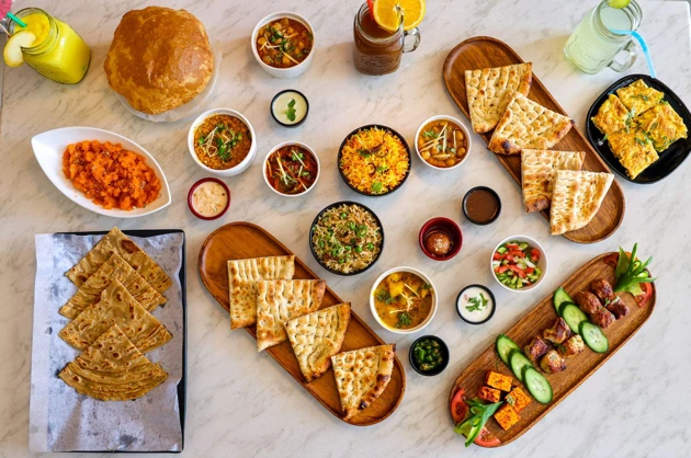 An image displaying authentic Pakistani cuisine, from Little Lahore, including Karahi, Tikka, Naan, Parathas, Puri, with chutneys and drinks over a white backdrop.