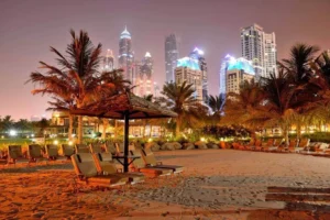 A night view of Jumeirah Beach in Dubai, with brightly lit skyscrapers in the background and the soft glow of lights along the beach, creating a lively combination of city and sea with buildings