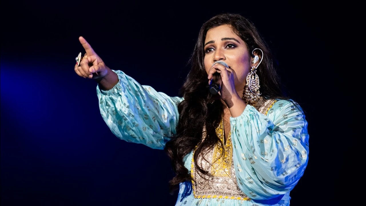 Versatile Shreya Ghoshal performing on stage under blue light, in a shimmering sky-blue gown, holding a microphone in one hand while pointing forward with the other.